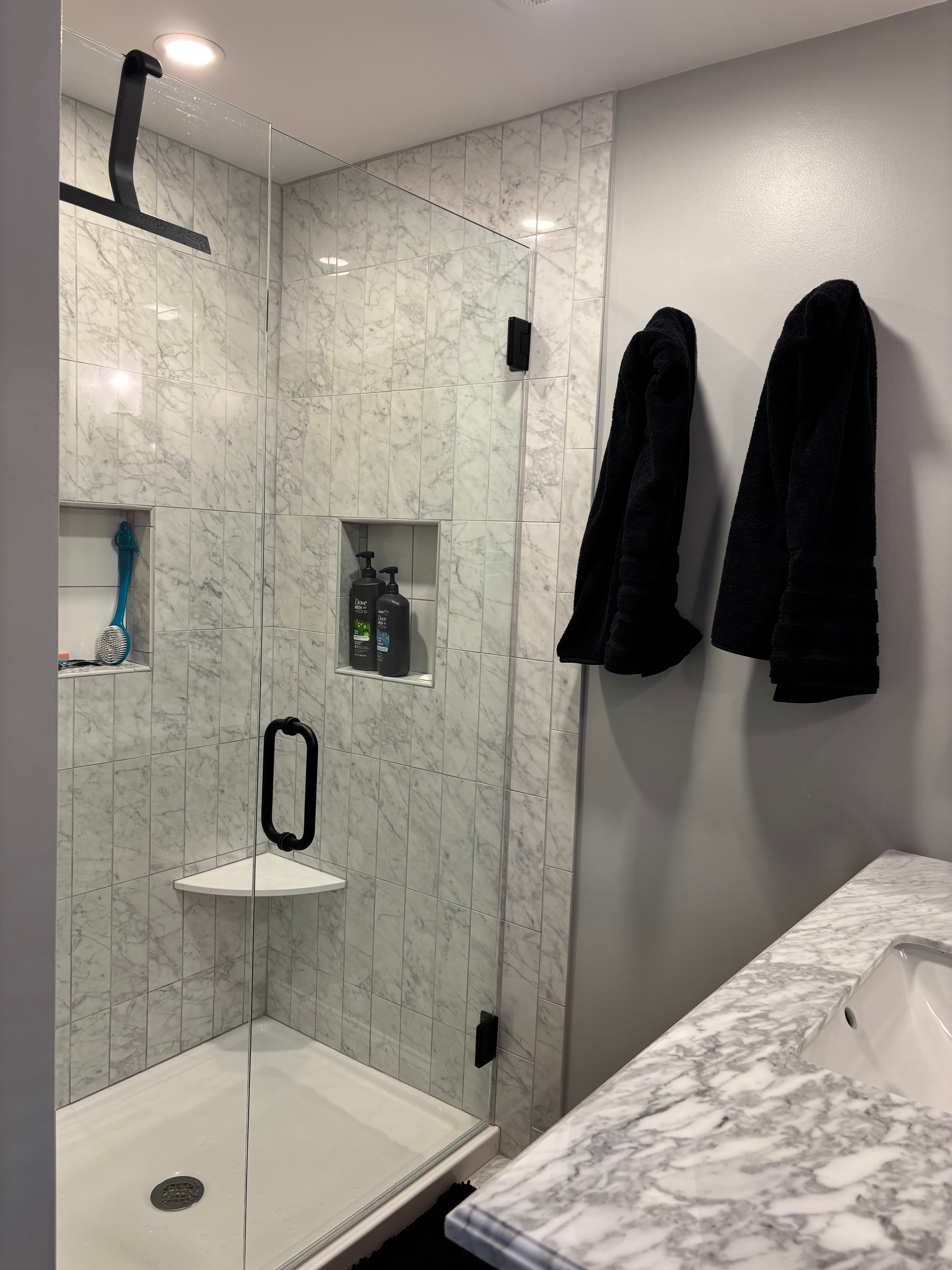 Completed Boston J's glass-enclosed walk-in shower with marble-look tile, black fixtures, two recessed niches and corner seat