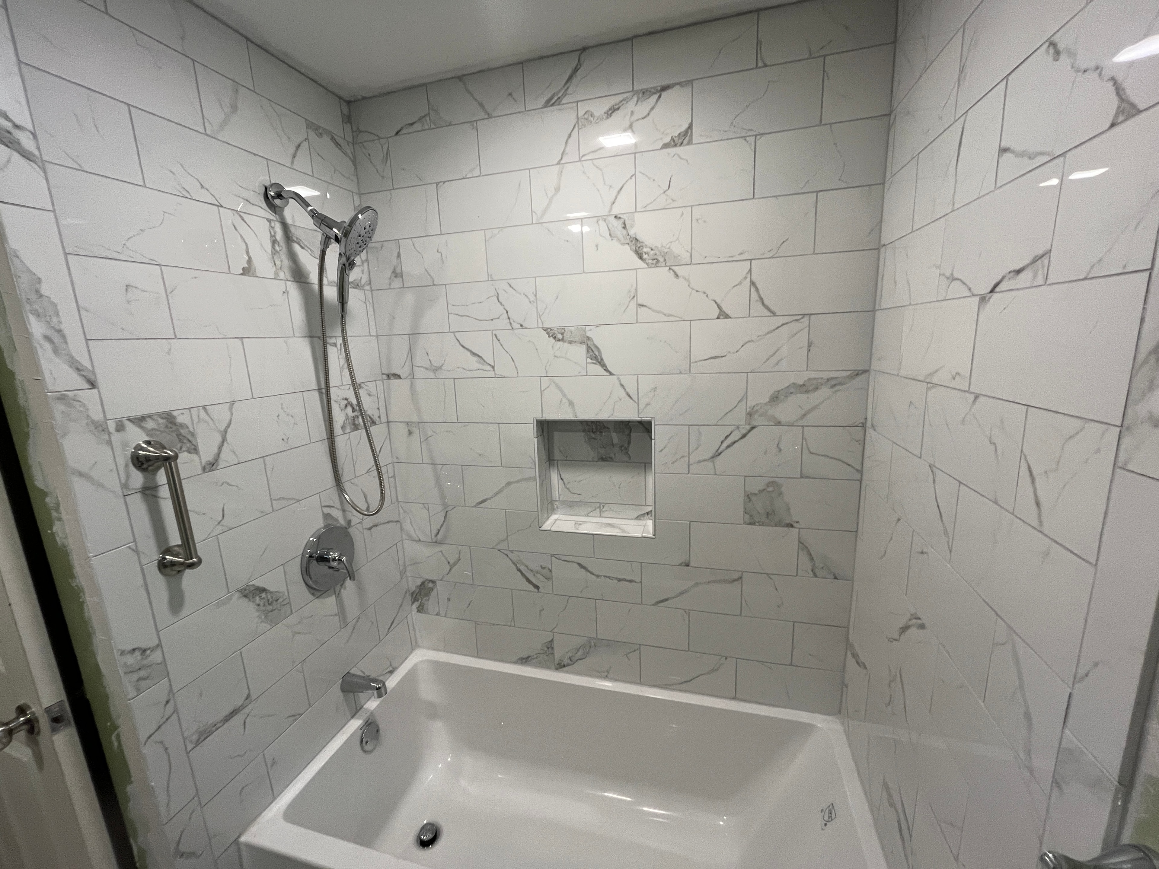 Completed Boston J's bathroom remodel showing marble-look tiled tub and shower surround with recessed niche, handheld shower and safety bar
