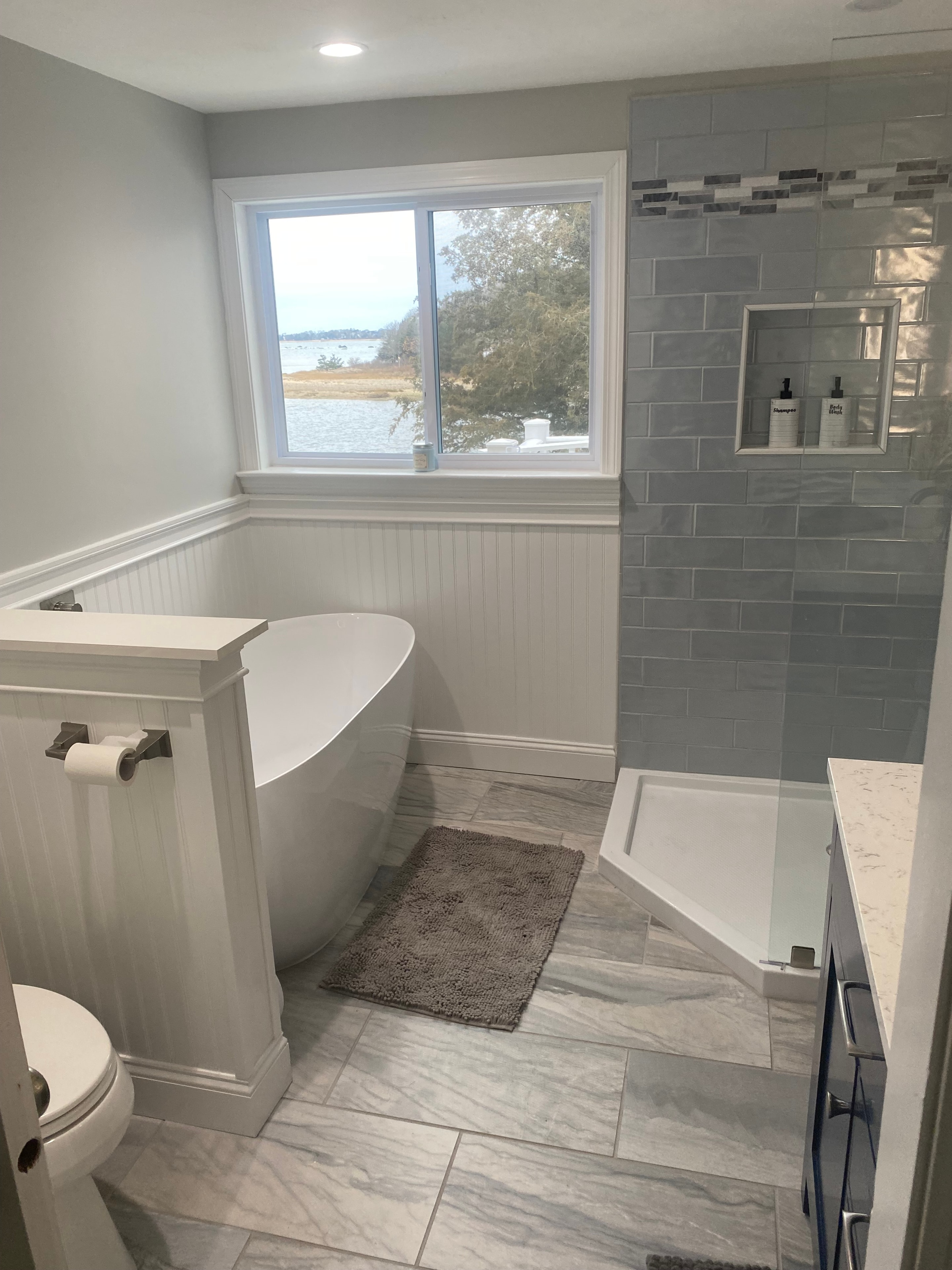 Completed Boston J's bathroom remodel showing freestanding bathtub, tiled shower surround, wainscoting and water view through window