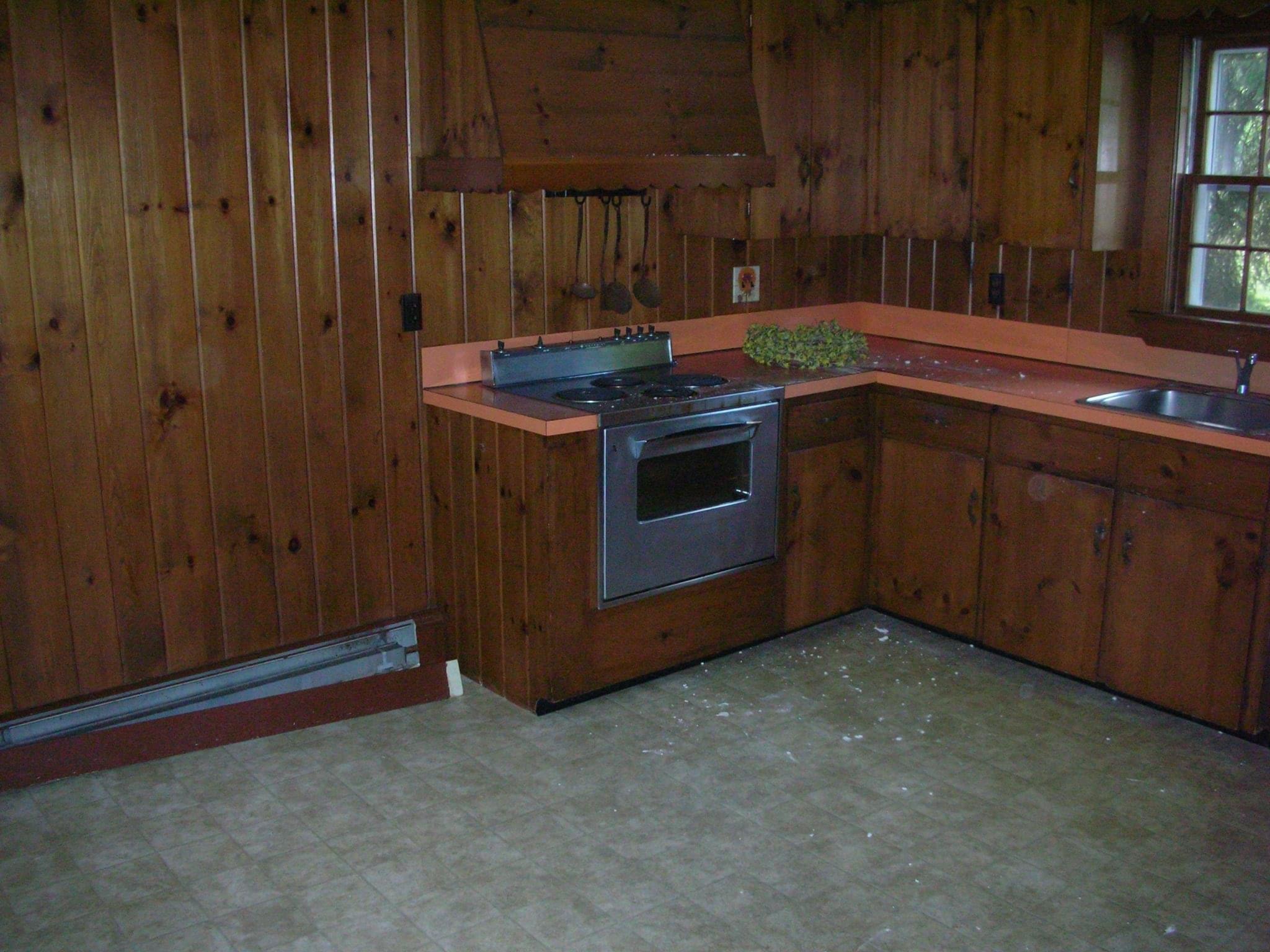 Before photo of a dark wood-paneled kitchen with older cabinetry and pink countertops prior to Boston J's remodel