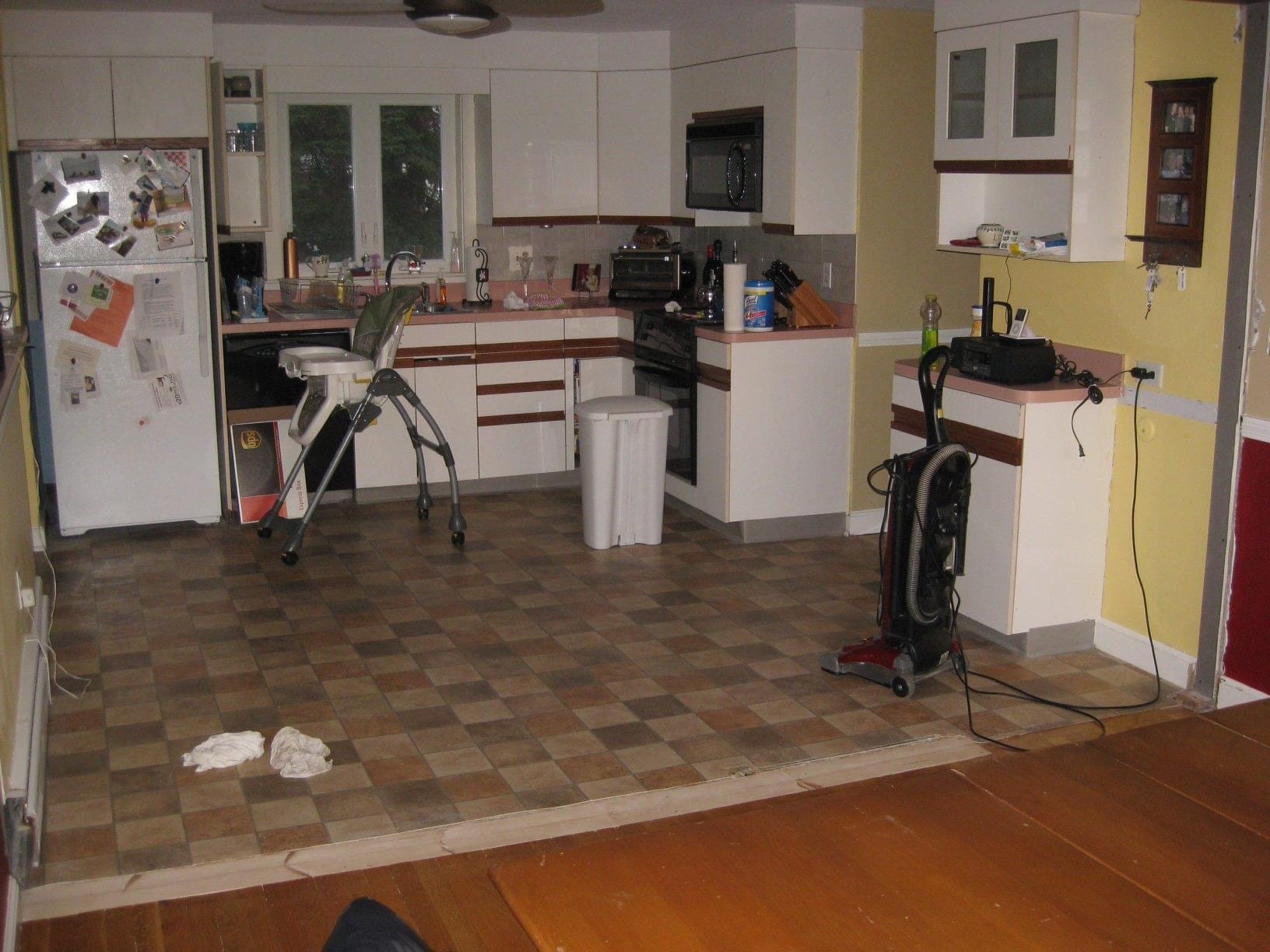 Before photo of a kitchen with white cabinets, dark horizontal trim, patterned flooring and yellow walls prior to Boston J's remodel