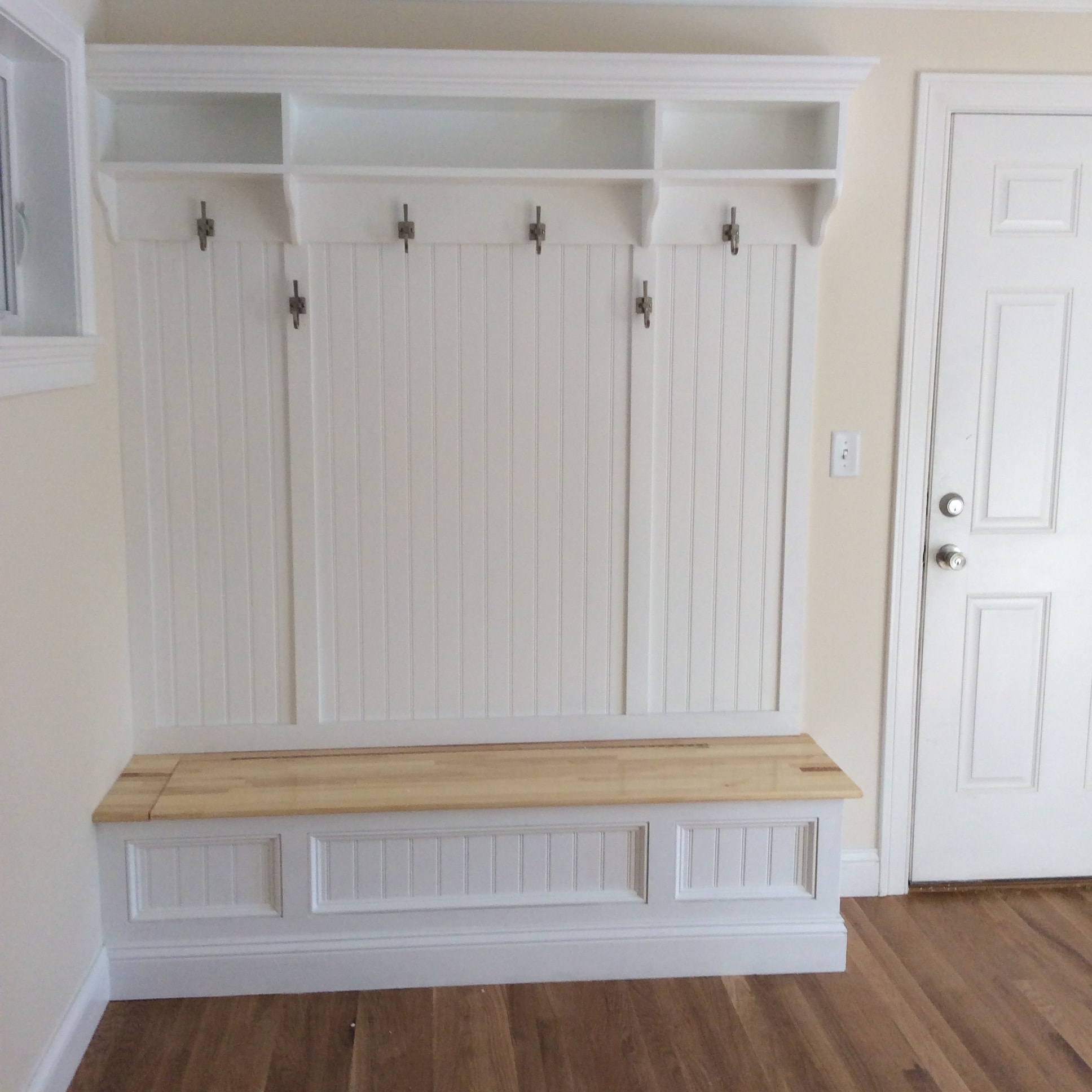 Custom entryway built-in by Boston J's featuring a fitted wood bench, beadboard wall treatment and coat hooks
